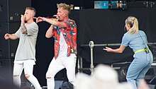 Members of L27, two white men, both wearing white pants, one wearing a red Hawaiian shirt and the other a gray t-shirt, sing into microphones on stage.
