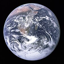 A planetary disk of white cloud formations, brown and green land masses, and dark blue oceans against a black background. The Arabian peninsula, Africa and Madagascar lie in the upper half of the disk, while Antarctica is at the bottom.