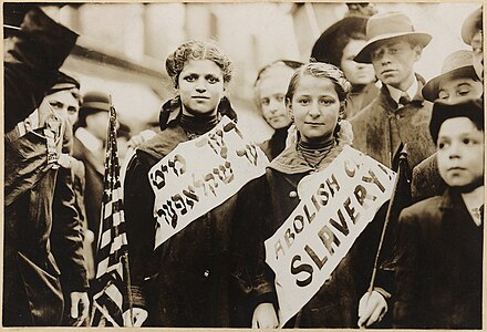 Üstünde İngilizce ve Yidiş dilinde "Çocuk Köleliğini Durdurun" sloganı yazılı olan bant taşıyan iki kız. Büyük olasılıkla 1 Mayıs 1909'daki New York işçi yürüyüşünde çekilmiş bir fotoğraf. (Üreten: Bain News Service photograph)