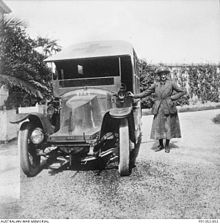 a black and white image of an early 20th century lorry, with a cross painted on the bonnet. Standing on the left of the vehicle is a woman, wearing a military uniform, with a long, below the knee skirt, a jacket belted at the waist, stockings, shoes, and a hat. She is smiling at the camera, with her left hand on her hip, and her right hand resting on the vehicle.