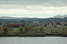 A collection of ruined farm buildings and a nissen hut sit amid fields that lie beyond the grey waters of a river. Wooded slopes and higher treeless hills lie beyond.