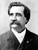 Black and white photo of a man with dark hair and an enormous moustache. He wears civilian garb: a dark suit and a white shirt.