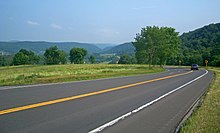 A paved two-lane road crosses the lower half of the picture from left to right. A small blue car is visible at the right with its brake lamps on, where the road turns back sharply. After the turn, it is slightly downhill from the camera and cannot be seen, although black-on-yellow arrow signs indicate where it is. In the distance, in the top of the image, is a valley with green hills on either side