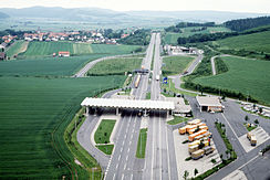 Vue aérienne d'une autoroute à quatre voie traversant les champs avec un petit village à l'arrière-gauche de l'autoroute. Au premier-plan se trouve une structure avec un toit blanc reposant sur quatre piliers blancs fins surplombant les quatre voies. À droite, on peut voir un parking avec des camions de couleur orange garés en diagonale.