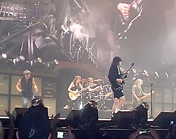 Rock band in performance on a well-lit but hazy stage. we see two guitarists, a bassist, a vocalist off to one side, and a drummer in the rear.