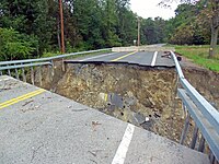 A road with a huge gap in front of the camera. Pieces of marked pavement are visible in the hole; its bottom cannot be seen from this angle. The box girder guardrails continue across it on either side. Beyond it is a concrete barrier