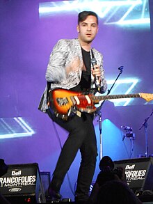 Blurry image of Dumas wearing black pants and a sparkly jacket, standing onstage with a guitar around his neck, holding a microphone