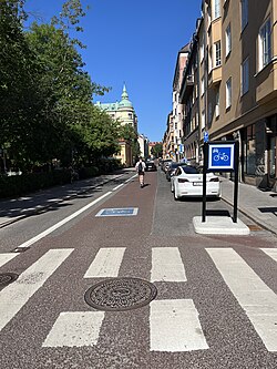 Cykelgata. Man ser en korsning och en cykelgateskylt. På gatan står bilar parkerade och en elsparkcyklist kör i mitten av vägen. Asfalten är svagt rödfärgad.