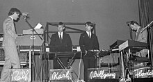 A black and white photograph of four members of Kraftwerk onstage, each with a synthesizer