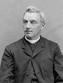 The upper-body of a middle-aged male looking slightly to his right. He wears the black robe and white collar of a Catholic priest
