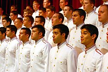 28 people standing on risers are signing while wearing white military-style uniforms