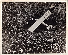 Black-and-white photograph of an enormous crowd of peoplem, packed like sardines, crowding aroud a small aircraft marked NX211 on the starboard wing