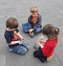 Trois enfants âgés d'environ six ans sont groupés assis sur le sol, un garçon et une fille agenouillés, l'autre garçon assis en tailleur. Les deux enfants agenouillés tiennent des billes ; il y a d'autres billes dans un sac sur le sol. Les trois enfants les regardent.