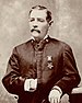 A white man with a mustache standing with his left arm resting on an object to his side and his right hand inside his jacket. A star-shaped medal is hanging from a ribbon on his left breast.