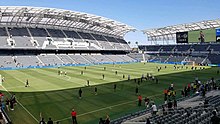 View of a soccer field from the stands of Banc of California Stadium with some players training on the field and a sparse number of fans in the stands. A third of the field is covered by shade.