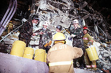 Plusieurs pompiers et membres de l'Armée de l'Air sont en train de nettoyer les débris de l'immeuble endommagé. Ils utilisent les seaux jaunes que l'on peut voir sur la photo pour y stocker les débris. Les dégâts causés par l'explosion sont visibles derrière les secouristes.