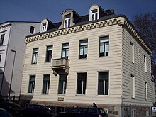 Light-coloured two-storey stone building with an ornamental ledge below the roof. There are rows of five windows at ground level and the floor above, with a balcony at the middle upper window.