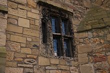 A wall of irregular stones, mostly light reddish brown, in which a large arch is set. This has been walled in, with a window in the centre, it also walled in, all in stone