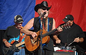 An old man wearing a black shirt, cowboy hat and sunglasses plays the guitar while also singing on the microphone, he is accompanied by a guitarist and a drummer in the background