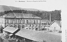 Photo noir et blanc montrant un bâtiment rectangulaire en pierre entouré de petits bâtiments en bois ou maçonnés.