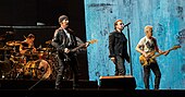 A color photograph of four men, two of which are playing the guitar. The third man is singing through a microphone.