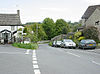 Street scene showing a road unction with parked cars. On the left and right are stone houses with trees and fields in the distance.