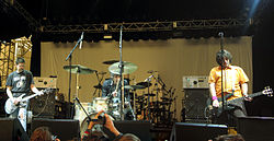 Color photo of three men playing on stage, with the guitarist at left, the drummer in the middle, and the bassist on the right.