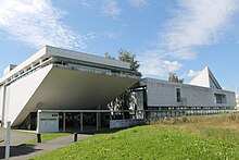 Frontale Farbfotografie eines Betongebäudes, aufgeteilt in zwei Komplexe. Links geht die vordere Mauer schräg nach hinten und hat oben eine schmale Fensterreihe sowie im Erdgeschoss einen Glaseingang. Rechts ist ein rechteckiger Gebäudeteil mit einem dreieckigen Aufbau, zwei unterschiedlich großen Fenstern und im Erdgeschoss eine Fensterreihe.