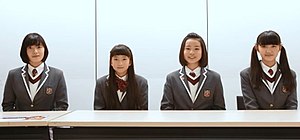 Four girls wearing school uniform outfits sit at a table against a white background