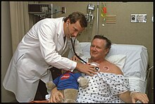 A view of a male doctor, wearing a white lab coat, using a stethoscope to check on the patient who was the 1,000th open-heart surgery patient at the Riverside Heart Institute of Ohio, the cardiovascular unit of Riverside Methodist Hospital. The male patient is holding a stuffed toy bear wearing a T-shirt that reads, "Riverside Heart Institute of Ohio."