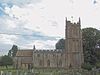 Stone building with arched windows. Prominent square tower to the right hand end. In the foreground are gravestones in grassy area.