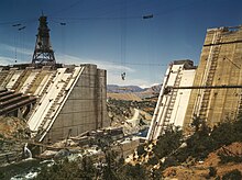 A crane lifts material used in the construction of a large concrete dam, which is still notched in the center to let the water flow through. Several pipes for future water conveyance use are visible on the left.