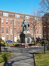 Statue on a plinth in park area, with buildings behind.