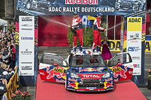 Sébastien Loeb et Daniel Elena sabrant le champagne debout sur le toit de leur Citroën DS3 WRC, portières ouvertes et entourée de spectateurs.