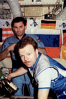 Two men seen in front of a wall featuring a number of switches and dials and covered by large American, Russian and German flags. The men are wearing blue jumpsuits, and two ventilation hoses are visible at the top of the image.