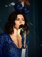 A young brunette woman singing into a microphone. She is wearing a blue sparkling dress and has a headpiece with a pair of blue sparkling cherries on it.