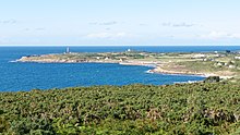 Photographie d'une pointe de terre avançant dans la mer, de la droite de l'image vers la gauche. Un phare se dresse à son extrémité.