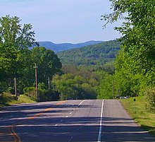 A four-lane highway descends a hill and disappears from view. In the background are large, tree-covered mountains.