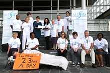 A group of 15 people posing in front of a building. One person lays on a cot holding a sign that reads, "I'm Here...23 hrs a day"