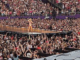 Taylor Swift stands on a stage surrounded by a large crowd of fans.