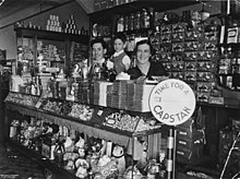 Stacks of chocolate bars line the top of the counter above an assortment of lollies in the glass cabinet. Ornaments and toys are on the lower shelf of the counter. Rows of tinned food, jars and cardboard boxes line the shelves behind the family. A large sign to the right of the counter advertises Capstan cigarettes.