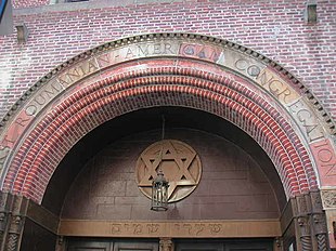 The top of an arched reddish-brick entrance-way is visible. Carved into stones on the top row of the arch are the words "First Roumanian-American Congregation", all in capital letters. The arch surmounts a brown wall with a bronze Star of David on it, with a lamp hanging from the arch in front of it. Underneath the brown wall, and above the doors, are inscribed the words "Shaarey Shamoyim" in Hebrew.