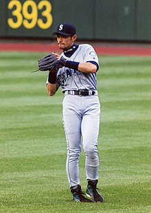 A Japanese man wearing a grey Seattle baseball uniform fielding a ball in the outfield.