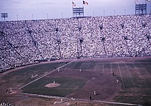 A baseball game being played in a stadium with high stands, seen from the right field side. The United States and Los Angeles flags are flying above the left field foul line, which is shorter than usual