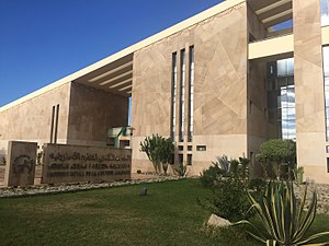 The entrance to the IRCAM (Institut Royal de la Culture Amazighe) building in Rabat. Two women are standing in front of a large, metal plaque with the word "IRCAM" written on it in large letters, and the organization's logo.