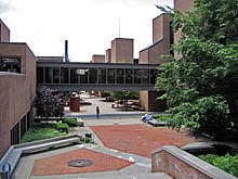 A brick courtyard is flanked by three-story brick buildings with a black glass bridge between them. Trees are visible to the right.