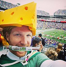 A man wearing a green Packers jersey and a large wedge of fake cheese on his head