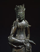 Photographie d'une sculpture d'un bouddha en métal. Il est assis, sa jambe gauche est ramenée sur le genou de sa jambe droite. Il est disposé dans un fond neutre.