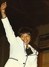 A photo of a woman wearing a black and white blazer, her hair in a bob, and a gold chain, raises her right fist in a black power salute, while smiling.