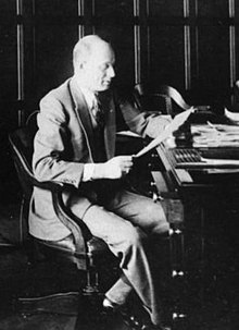 A black-and-white, two-thirds profile photo of a man wearing a suit, sitting at a desk, and looking at a piece of paper.